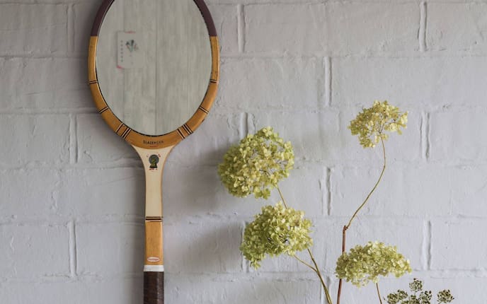 Alter Holztennisschläger von Slazenger hängt an einer weiß gestrichenen Backsteinwand. Dort wo das Netz des Schlägers war ist jetzt ein Spiegel angebracht.