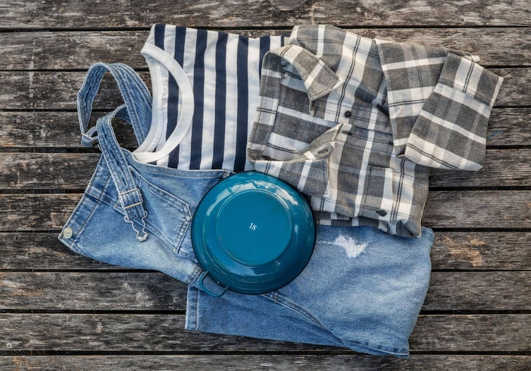 Auf einem Holzuntergrund liegt die Kleidung der Vogelscheuche: eine Latzhose aus Jeans, ein blau-weiß gestreiftes T-Shirt, ein grau-weiß kariertes Flanellhemd, ein blauer Emailletopf.