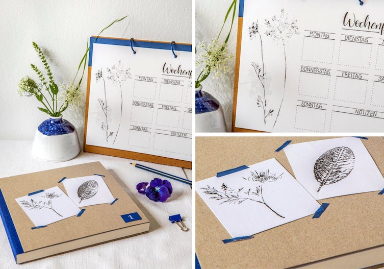 Ein selbstgebastelter Wochenplan-Stehkalender mit Linoldruck von Blüten und Blättern. Daneben liegt ein Buch mit braunem Kartondeckeln. Darauf sind zwei kleinere quadratische weiße Papierstücke mit blauem Klebeband befestigt, die auch einen Linoldruck von Blättern und Blüten haben.
