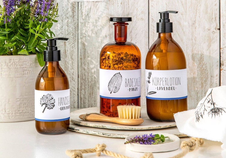 Braune Apothekerflaschen mit Glasstoppel oder schwarzen Kunststoffpumpen sind mit weißen Etiketten, die am unteren Rand blau eingefärbt sind, mit einem schwarzen Stift in Schönschrift beschriftet (Handseife Geranium, Badesalz Minze, Körperlotion Lavendel). Neben dem Schriftzug ist ein Blatt im Linoldruck gedruckt. Passend zum verwendeten Kraut.