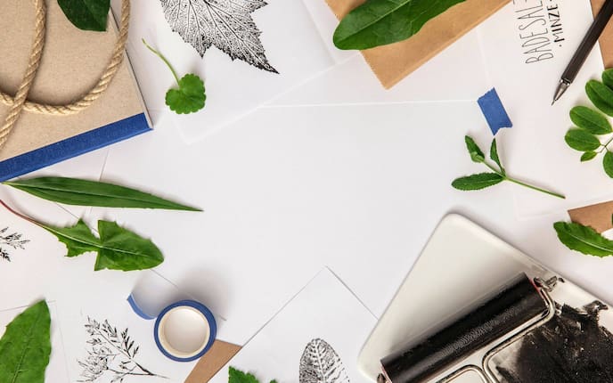 Weiße Unterlage von oben fotografiert. Es liegen Blätter herum, eine Linolwalze mit schwarzer Farbe, weiße Blätter, Backpapier, ein blause Klebeband, ein Druckbleistift, eine Ecke von einem Buch ist zu sehen, ein dünnes Sisalseil.