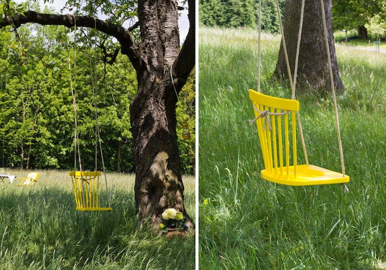 Sommerwiese mit alten Bäumen, auf einem Baum ist ein gelber Holzsessel auf Seilen auf einem Ast aufghängt.