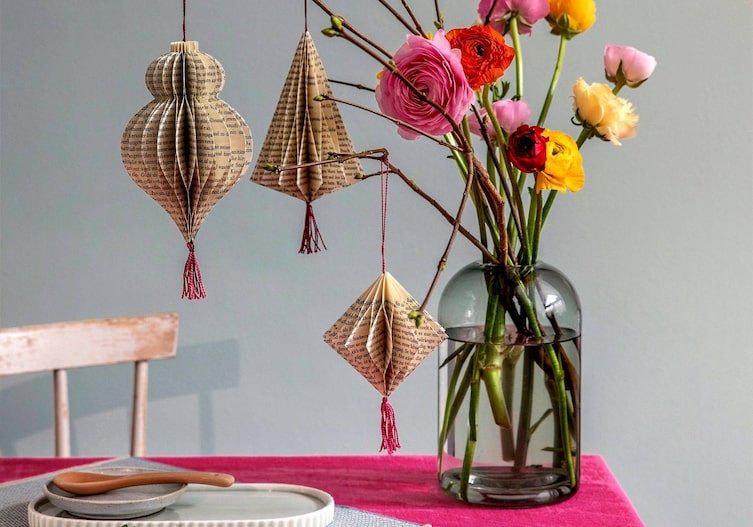 Ranunkeln und Rosen und Zweige stecken in einer Glasvase. An den Zweigen hängen 3D-Ornamente aus Buchseiten gefaltet