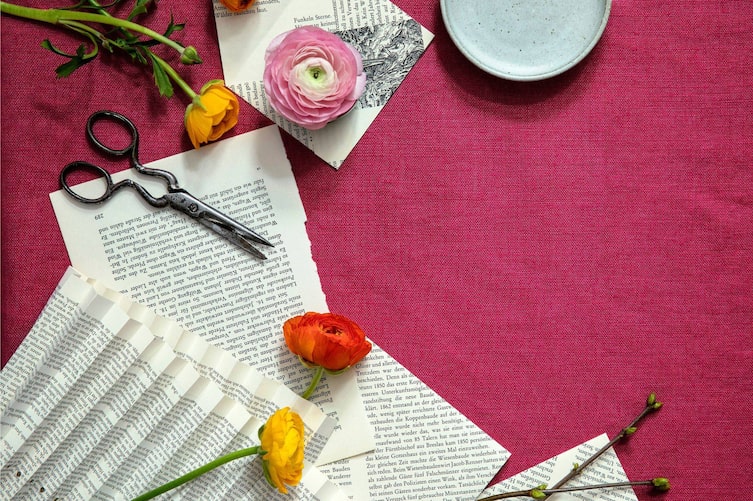 rote Tischtuch, darauf liegen Ranunkeln, eine alte Schere und alte Buchseiten
