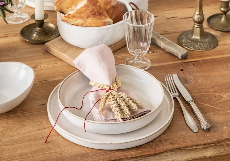 Ein Holztisch mit schlichtem weißen Gedeck darauf liegt eine zartrosa Stoffserviette in einem geflochtenem Ährenring.
