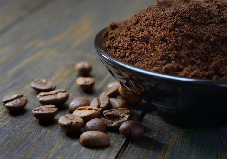 Kaffeepulver in einer dunklen Schale, daneben liegen Kaffeebohnen auf einem Holztisch