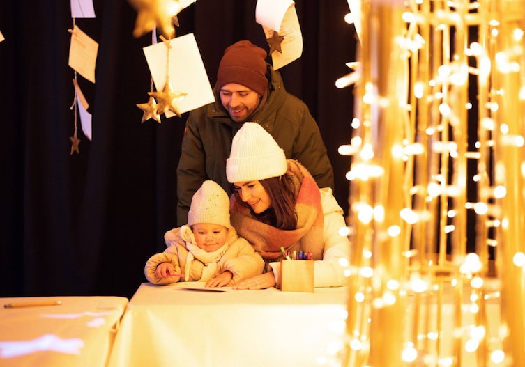 Vater, Mutter und Kleinkind beugen sich gemeinsam über ein Blatt Papier an einem Basteltisch, umgeben von goldenen Lichterketten und hängenden Sternen
