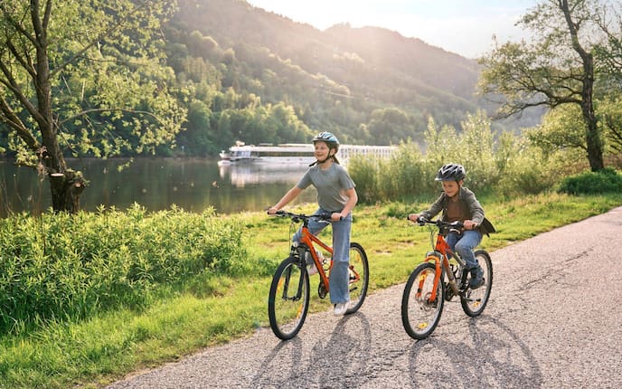 Zwei Kinder mit orangefarbenen Fahrrädern und Helmen auf einem Weg entlang der Donau, im Hintergrund ein Flussschiff und bewaldete Hügel