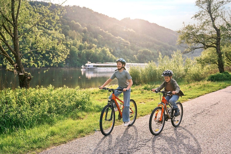 Zwei Kinder mit orangefarbenen Fahrrädern und Helmen auf einem Weg entlang der Donau, im Hintergrund ein Flussschiff und bewaldete Hügel
