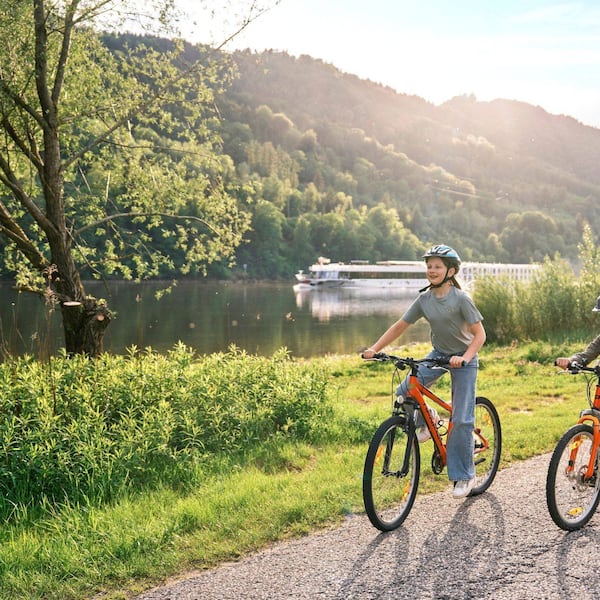 Zwei Kinder mit orangefarbenen Fahrrädern und Helmen auf einem Weg entlang der Donau, im Hintergrund ein Flussschiff und bewaldete Hügel