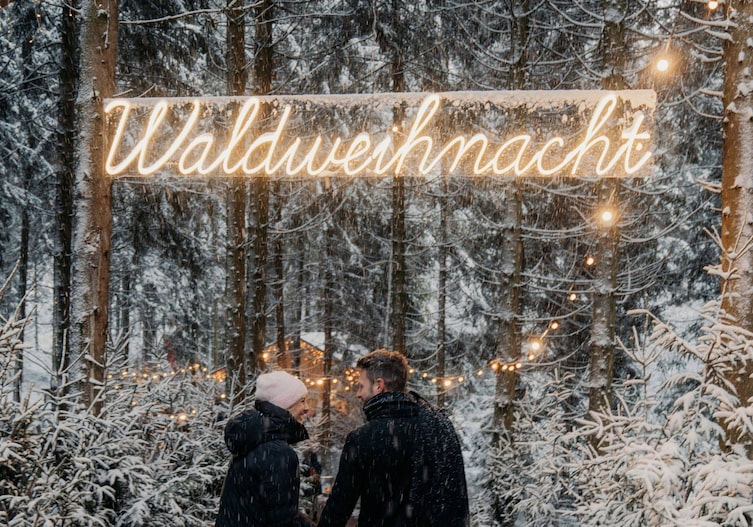 Zwei Personen im Schneetreiben vor einem leuchtenden Neonschild mit der Aufschrift „Waldweihnacht" in einem verschneiten Nadelwald