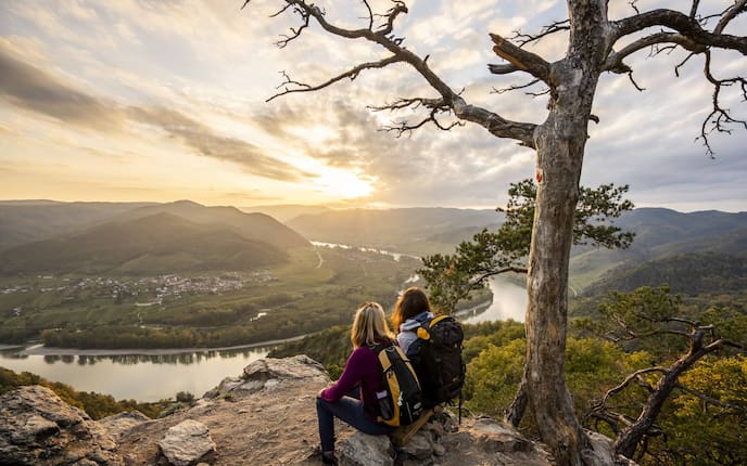 Zwei Wanderinnen sitzen auf einem Fels und blicken auf die Donau und eine grüne Hügellandschaft. Die Sonne geht gerade unter.