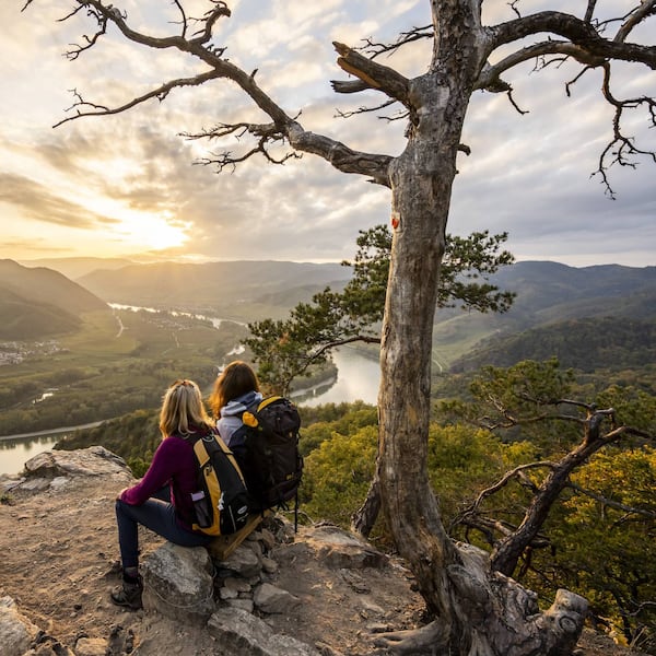 Zwei Wanderinnen sitzen auf einem Fels und blicken auf die Donau und eine grüne Hügellandschaft. Die Sonne geht gerade unter.