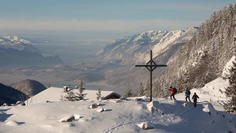 Drei Winterwanderer auf einem verschneiten Pfad in den Tiroler Alpen, vorbei an einem Gipfelkreuz, mit einem atemberaubenden Blick auf das weite Tal und die umliegenden schneebedeckten Berge.