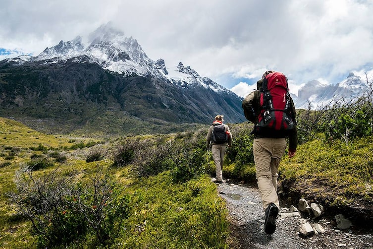 Zwei Personen wandern einen Weg entlang, vor ihnen befindet sich ein verschneiter Berg.