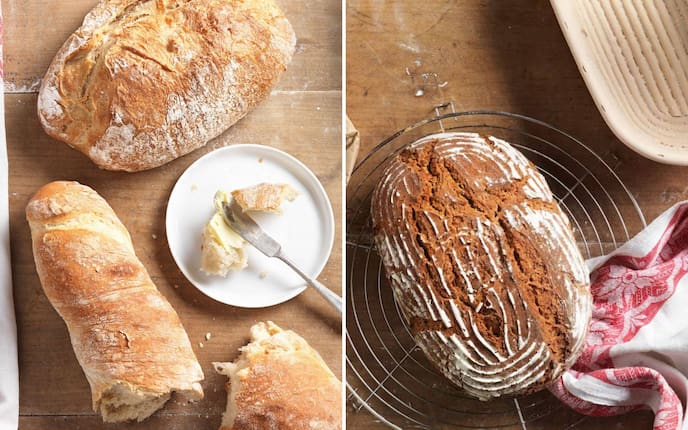 Draufsicht auf ein bemehltes Holzbrett mit einem runden, goldbraun gebackenen Weizenlaib und einem aufgebrochenen Baguette. Auf einem kleinen weißen Teller liegt ein Buttermesser mit etwas Butter, links ein weißes Küchentuch. Daneben: Rundlaib mit dunkler, rilliger Kruste auf einem runden Drahtgitter-Abkühlrost auf einem Holztisch. Das Muster stammt von einem Gärkorb, der im Hintergrund zu sehen ist. Ein rot-weißes Küchentuch ist um den Laib gelegt.