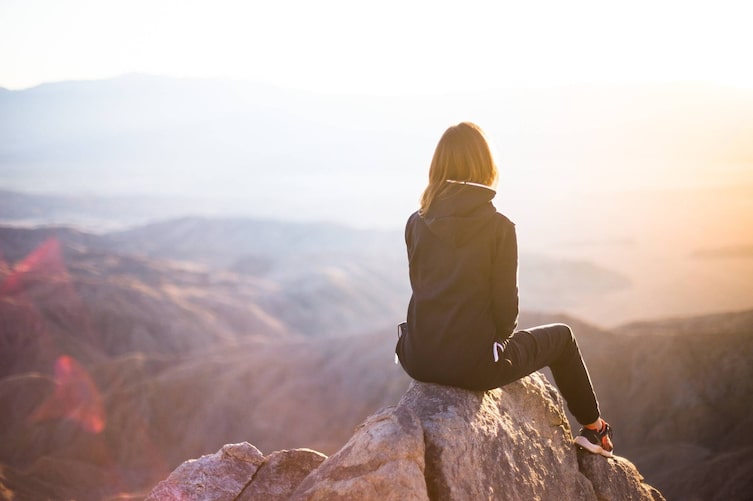 Frau von hinten sitzt alleine auf einem Fels und blickt über ein Bergpanorama
