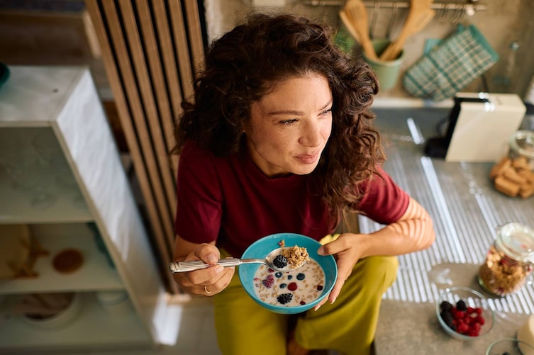 Eine Person isst ein gesundes Frühstück mit Müsli, Beeren und Milch in einer modernen Küche.