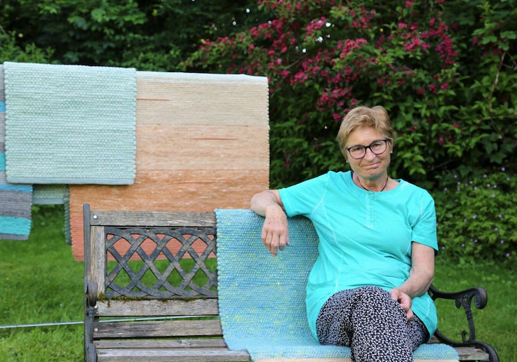 Christine Sickinger sitzt auf einer Bank mit ihren handgewebten Teppichen.