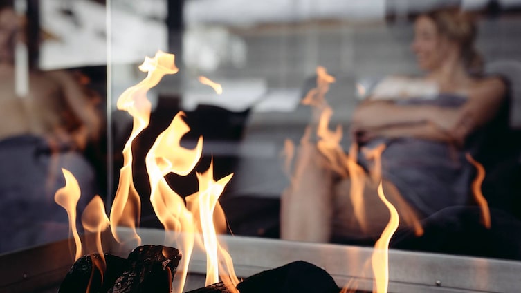 Ein Kaminfeuer flackert in einer Wellnessoase, im Hintergrund entspannt eine Person in einem gemütlichen Raum.