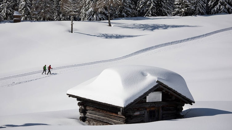 Zwei Personen auf einer Winterwanderung in Kartitsch, vorbei an einer tief verschneiten Holzhütte, umgeben von unberührter Natur und schneebedeckten Bäumen.
