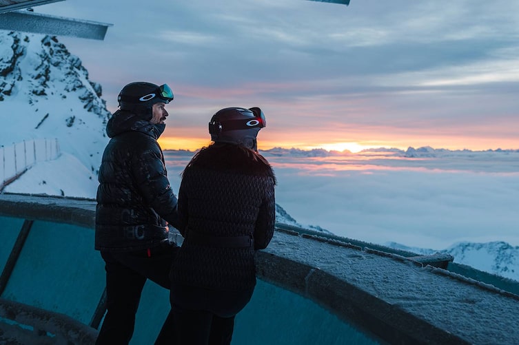Zwei Personen in Skibekleidung stehen an einer Aussichtsplattform hoch in den Alpen und blicken auf ein atemberaubendes Meer aus Wolken bei Sonnenuntergang.