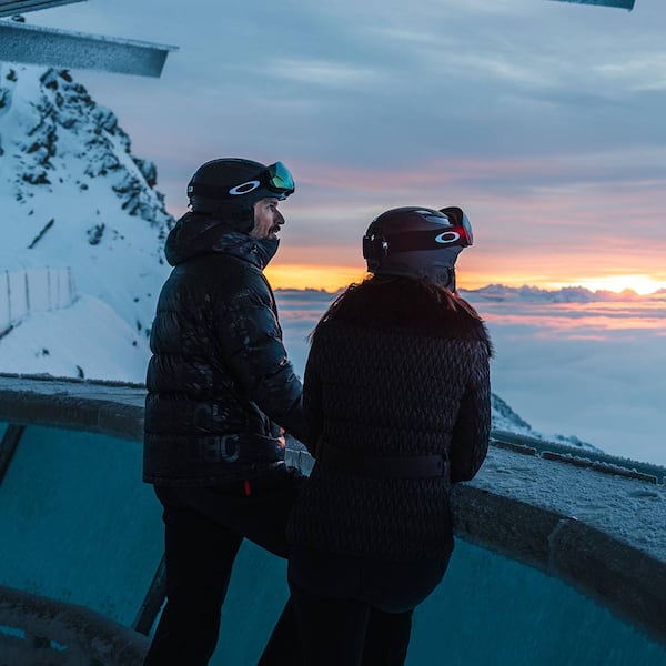 Zwei Personen in Skibekleidung stehen an einer Aussichtsplattform hoch in den Alpen und blicken auf ein atemberaubendes Meer aus Wolken bei Sonnenuntergang.