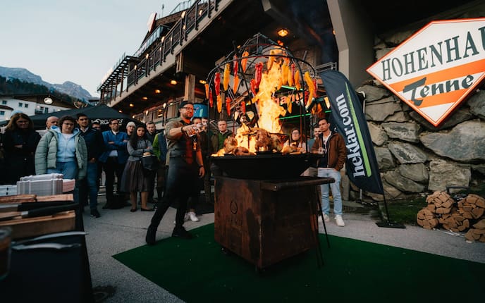 Ein Mann vor einem großen Griller mit Feuer und Grillhendl. Rund um ihn stehen Menschen und schauen zu.