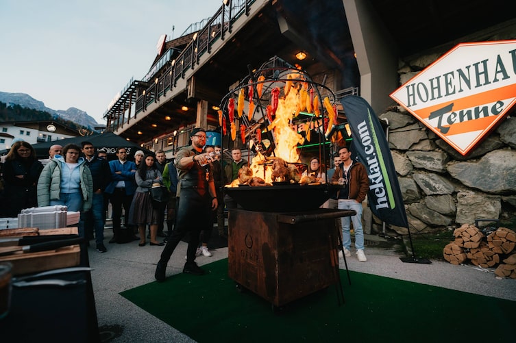 Ein Mann vor einem großen Griller mit Feuer und Grillhendl. Rund um ihn stehen Menschen und schauen zu.