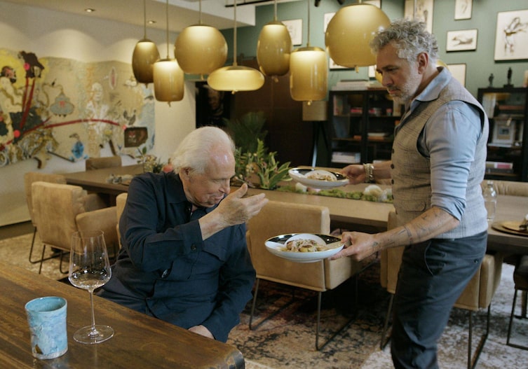 Zwei Männer, einer sitzt am Tisch, der andere bringt zwei Teller mit Fleischlaibchen und Gurkensalat zum Tisch und will sie servieren