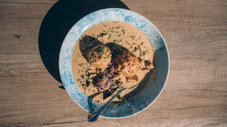 Ein Teller mit zwei fluffigen Kaspressknödeln in einer cremigen Pilzsoße, dekoriert mit frischen Kräutern und Sprossen, auf einem Holztisch serviert.