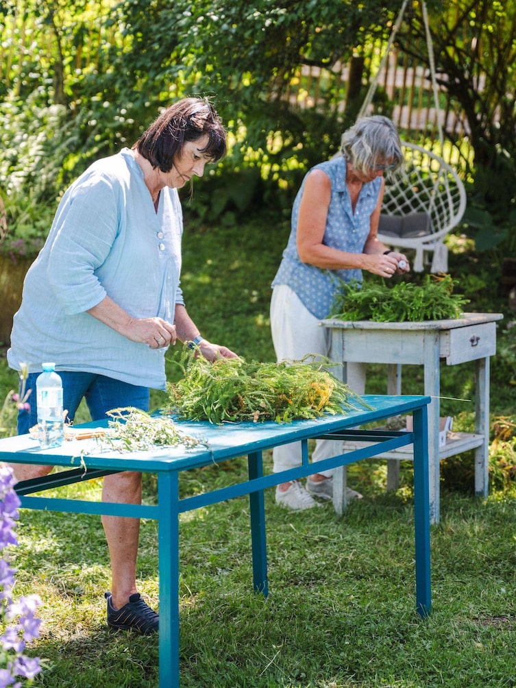 Zwei Frauen arbeiten an Tischen mitten im Garten an ihren Blumenkränzen.