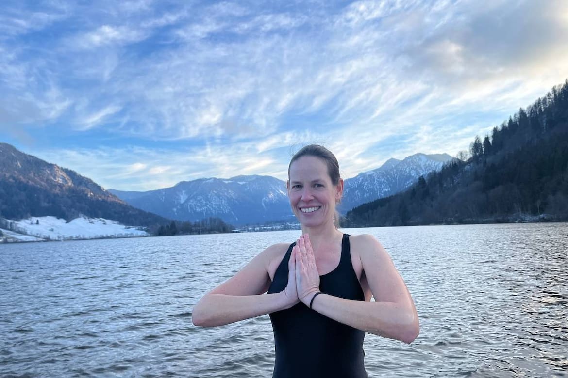 Iris Zenglein, Expertin zum Thema Eisbaden, steht in Yogapose vor See mit Bergpanorama