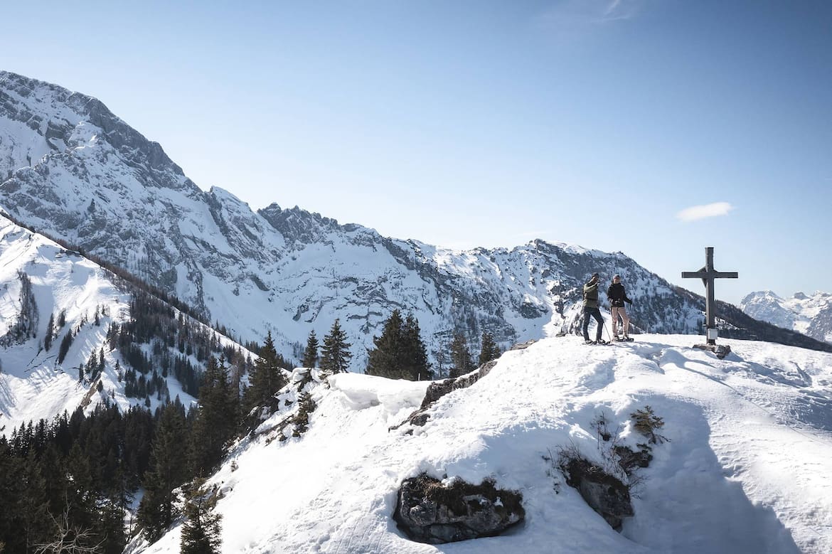 Eine Frau und ein Mann gehen Schneeschuhwandern und sind beim Gipfelkreuz angekommen.