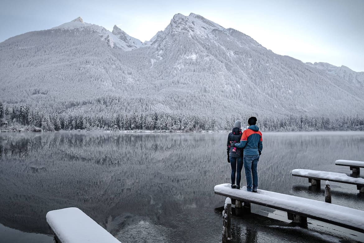 Eine Frau und ein Mann stehen auf einem Steg. Ihnen eröffnet sich ein alpines Panorama vor einem See.