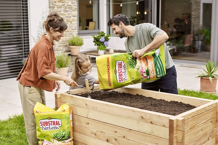 Eine Frau, ein Mann und ein Kind befüllen zusammen ein Holzbeet im Garten mit Erde.
