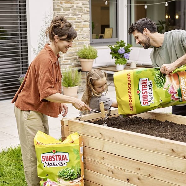 Eine Frau, ein Mann und ein Kind befüllen zusammen ein Holzbeet im Garten mit Erde.