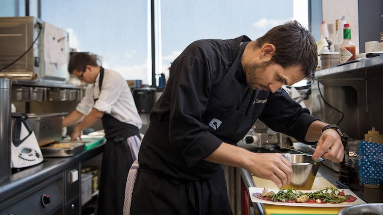 Ein Koch verfeinert mit Hingabe ein Gericht in einer modernen Küche, während im Hintergrund ein weiterer Koch sorgfältig Speisen vorbereitet – Tiroler Kulinarik mit Liebe zum Detail.