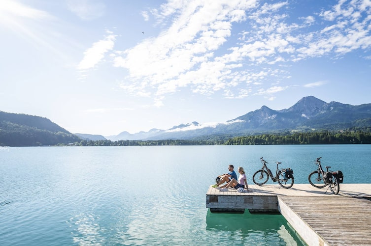 Eine Frau und ein Mann sitzen am Steg mit ihren Fahrradhelmen an ihrer Seite. Neben ihnen stehen ihre Fahrräder und sie blicken auf den Wörthersee.