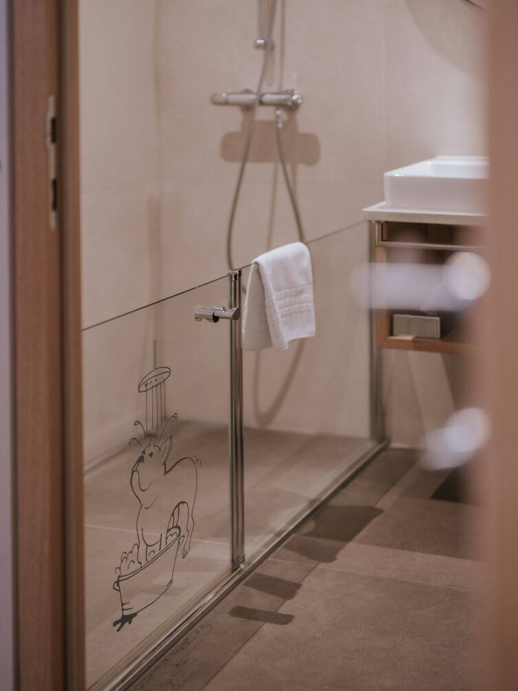 Hundedusche in einem Hotelzimmer, die Dusche ist mit einer halbhohen Glaswand vom restlichen Badezimmer abgegerenzt. Auf der Scheibe ist ein Hund abgebildet, der unter einer Dusche steht.