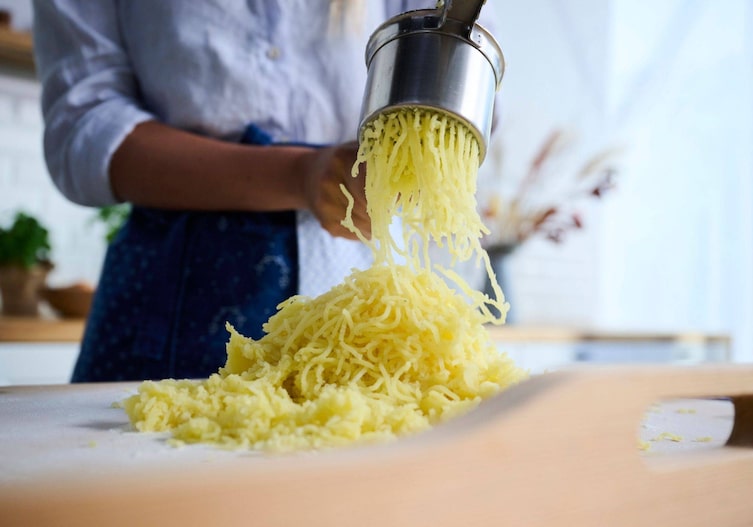 Gekochte Erdäpfel werden durch eine Kartoffelpresse auf eine Arbeitsfläche gedrückt.