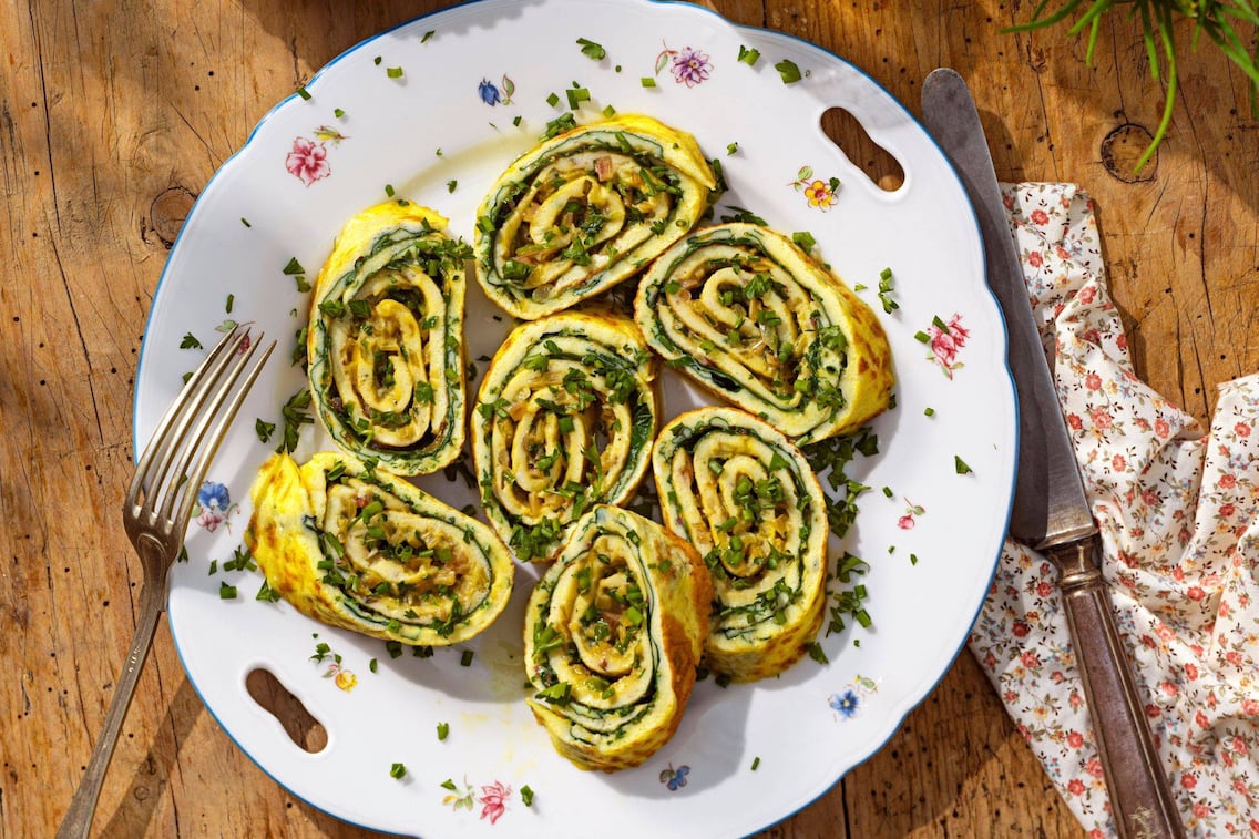 Omelette mit Mangold gefüllt in dünne Scheibchen geschnitten auf ienem Blümchenteller, mit Gabel und Messer und einen blumigen Stoffserviette. Alles zusammen ist von oben fotografiert und steht auf einem urigen Holzuntergrund.