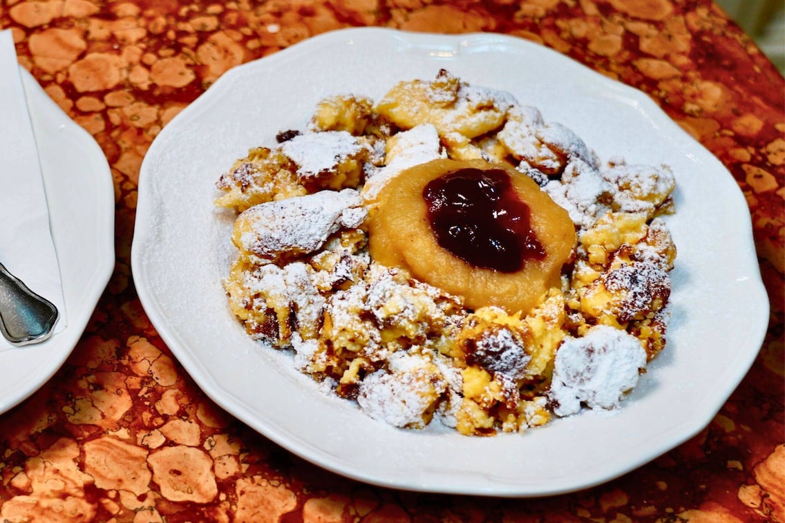 Kaiserschmarrn mit Staubzucker auf weißem Teller, in der Mitte Apfelmus mit dunklem Zwetschkenröster, serviert auf einer rotgemusterten Tischdecke. Ein Rezept von Paula Bründl.