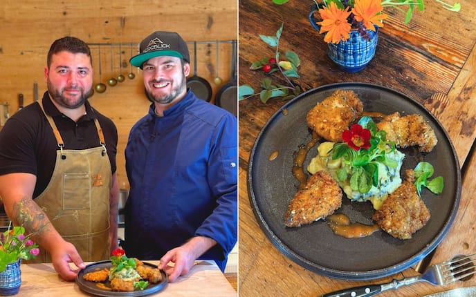 Andreas Ziep und Maximilian Nicolic in einer holzernen Küche mit ihrem Backhendl. Das wird präsentiert auf einem dunklen Teller mit einem Erdäpfelsalat und Vogerlsalat und einer Blume oben drauf.