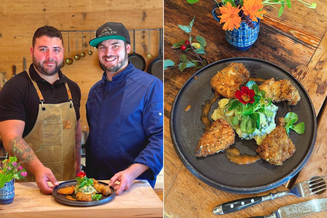 Andreas Ziep und Maximilian Nicolic in einer holzernen Küche mit ihrem Backhendl. Das wird präsentiert auf einem dunklen Teller mit einem Erdäpfelsalat und Vogerlsalat und einer Blume oben drauf.