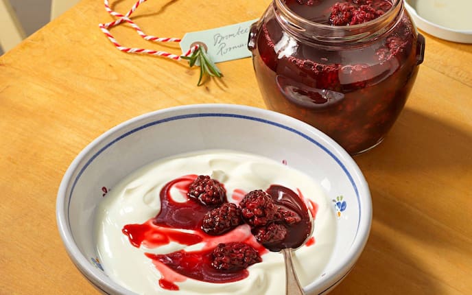 Eine kleine Keramikschüssel mit blauem Rand und kleinen bunten Blümchen, gefüllt mit Joghurt und Brombeerkompott. Danaben steht ein Einmachglas mit dem Kompott. In der Schüssel liegt noch ein Löffel. Alles steht auf einer Holzplatte.