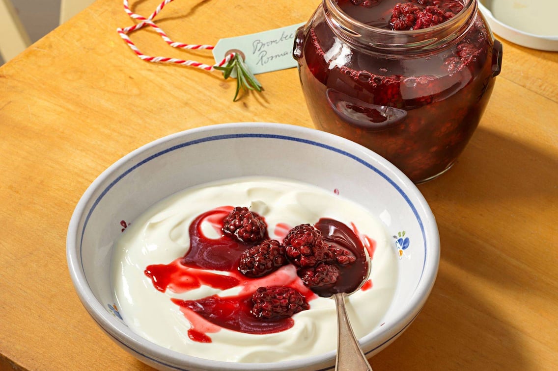 Eine kleine Keramikschüssel mit blauem Rand und kleinen bunten Blümchen, gefüllt mit Joghurt und Brombeerkompott. Danaben steht ein Einmachglas mit dem Kompott. In der Schüssel liegt noch ein Löffel. Alles steht auf einer Holzplatte.