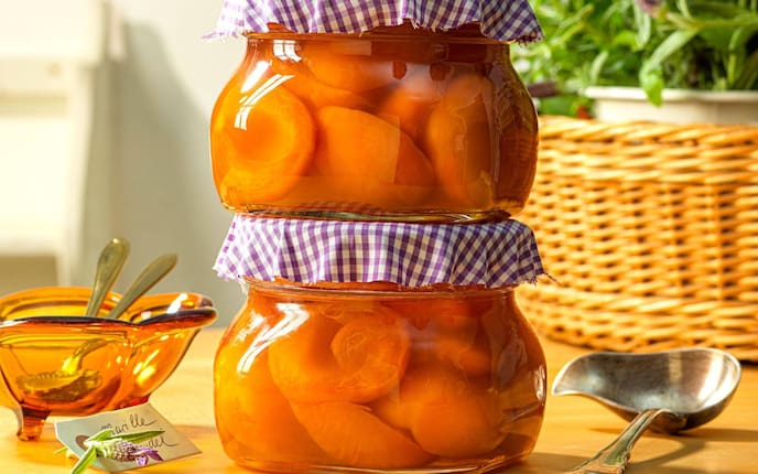 Marillenhälften zwei aufeinandergestapelten Einmachgläsern mit Stoffdeckel (violett kariert). Daneben liegt eine alte Suppenkelle ein orange, geschwungene Glasschüssel mit Löfferl. Dahinter steht verschwommen ein großer Korb mit Lavendel.