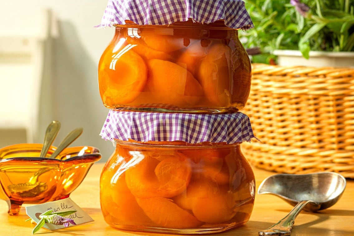 Marillenhälften zwei aufeinandergestapelten Einmachgläsern mit Stoffdeckel (violett kariert). Daneben liegt eine alte Suppenkelle ein orange, geschwungene Glasschüssel mit Löfferl. Dahinter steht verschwommen ein großer Korb mit Lavendel.