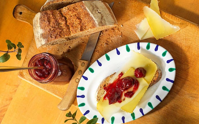 Auf einem Holzbrett steht ein halber Brotlaib ein offenen Kirschmarlmeladeglas mit einem Löffel drin, danaben steht ein ovals Gmundner Teller mit einem Stück Brot, Käsescheiben und Kirschmarmelade oben drauf.